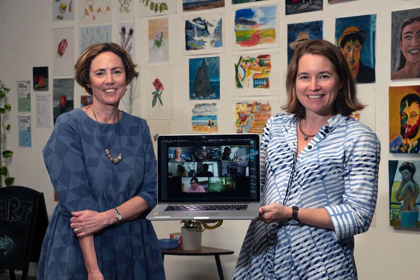 Two women pictured inside an art space holding a laptop with pictures behind them on a wall.